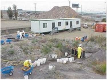 Photo showing excavation of the Emeryville Shellmound. Five people are working over blue wheelbarrows filled with material from the site. An office trailer sits in the background.