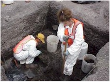 Two archaeologists work in an excavated hole. They are wearing white, with orange safety vests. The worker on the right is digging a hole with a shovel