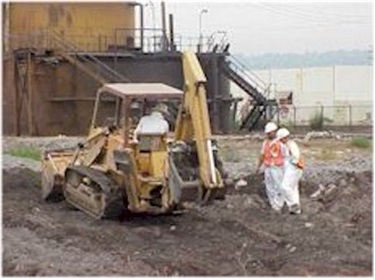 Photo of the Emeryville Shellmound excavation. A worker uses a backhoe while two other workers look on. An industrial building is visible in the background
