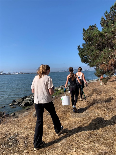 Three students walking away from the camera picking up trash along the Emeryville shoreline. The San Francisco Bay is to the left of the student.